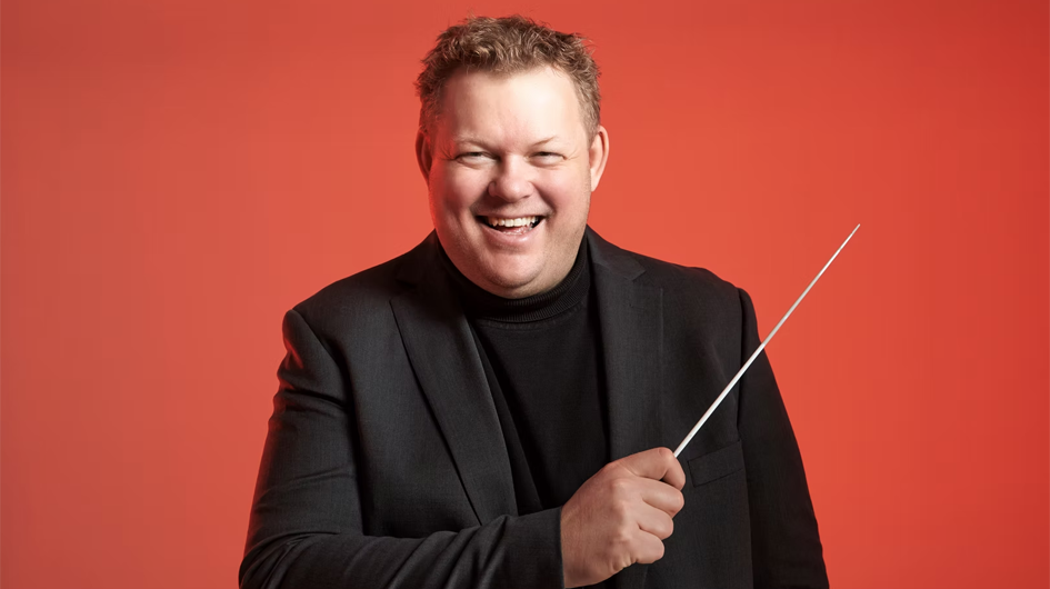 A promotional portrait of conductor Rune Bergmann, captured mid-laugh with a warm, open expression against a vivid coral-red background. He holds a conductor's baton in his right hand and wears a black turtleneck under a black blazer. The bright, energetic backdrop and candid joy of the pose give the image a modern, approachable feel.