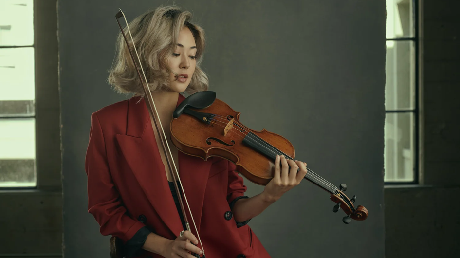 A cinematic promotional portrait of violinist Simone Porter, captured mid-performance in a dramatically lit, gray-walled interior space with tall windows on either side. She plays her violin with downcast eyes and quiet intensity, bow raised, wearing a vibrant red blazer over a dark top. Her short blonde hair frames her face softly. The warm, honey-toned wood of the violin contrasts vividly with both the red jacket and the cool, muted gray of the background, giving the image a bold, editorial quality.