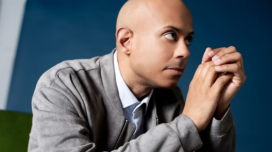 A contemplative promotional portrait of pianist Stewart Goodyear, shot against a rich deep blue background with a hint of green in the lower left corner. He leans forward with his hands clasped together beneath his chin, gazing upward and to the side with a pensive, focused expression. He wears a light gray suede zip-up jacket over a pale blue collared shirt. The shallow depth of field and close framing give the image an intimate, introspective quality. Photo credit: Anita Zvonar.