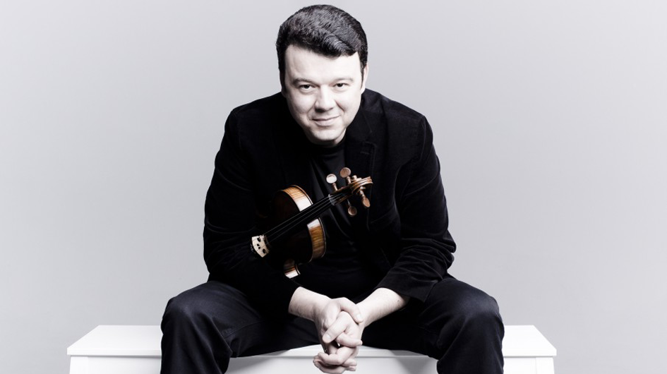 A clean, modern promotional portrait of violinist Vadim Gluzman seated on a white surface against a neutral gray background. He holds his violin tucked casually under his left arm, with his hands clasped together in front of him, leaning slightly forward in a relaxed pose. He wears an all-black velvet blazer and trousers and looks directly into the camera with a calm, confident expression. The warm wood tones of the violin stand out against the monochromatic styling of the image.