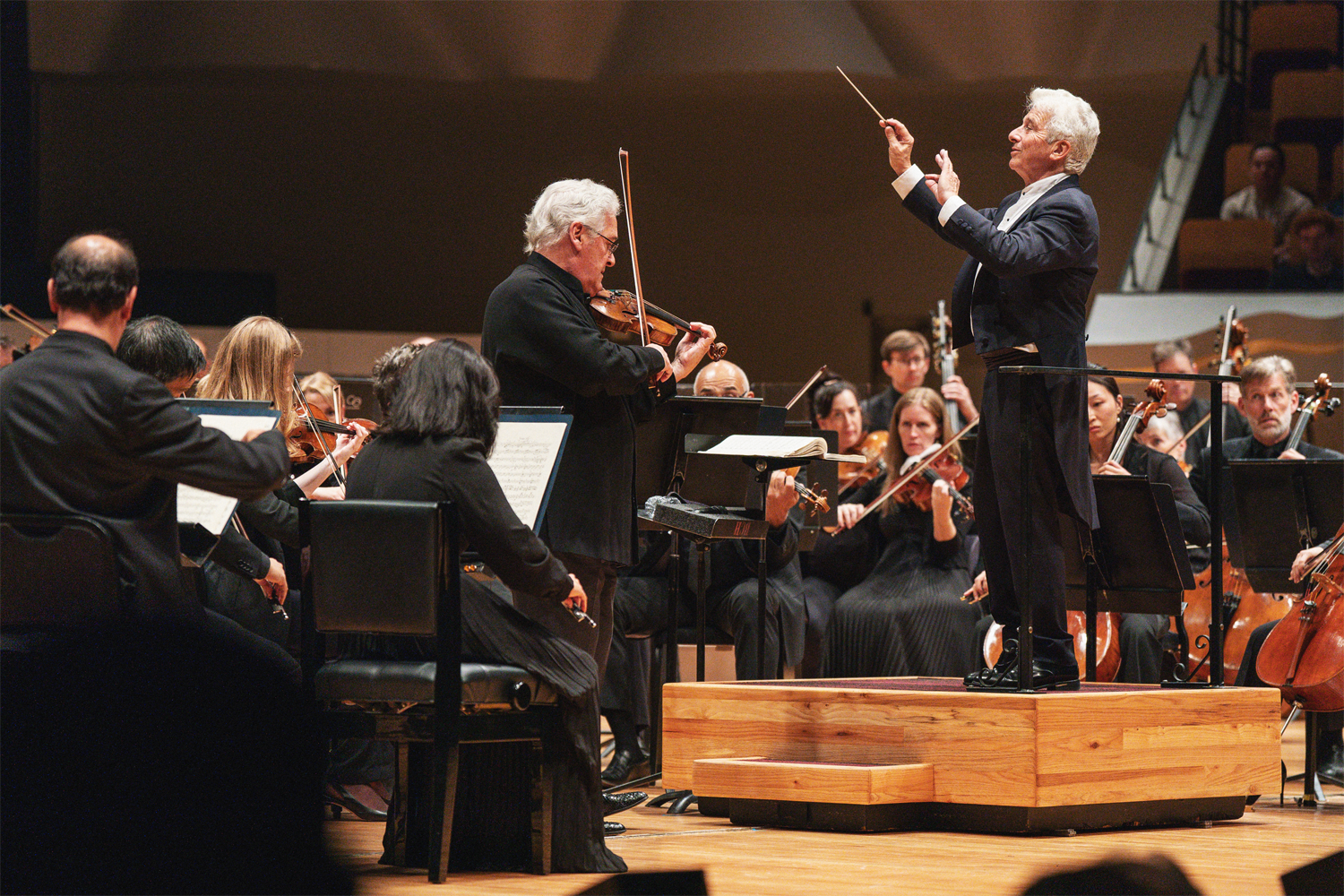 Violinist Pinchas Zukerman stands center stage at Boettcher Concert Hall, playing his violin with focused intensity while Music Director Peter Oundjian conducts expressively behind him from an elevated wooden podium, baton raised high and one hand extended outward. Both men have silver hair and wear formal black concert attire. The full orchestra surrounds them — string players with bows in motion are visible on both sides and in the background — creating a layered, dynamic image of a live classical performance in full flight. The warm wood tones of the stage floor and podium anchor the scene. Credit: Amanda Tipton Photography