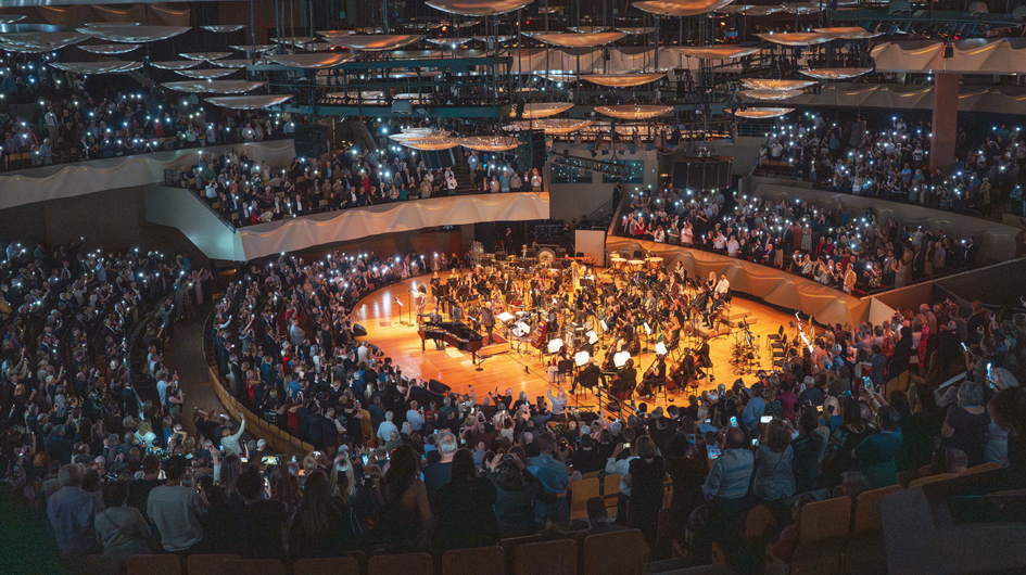 A breathtaking wide-angle overhead view of Boettcher Concert Hall completely filled with a standing audience, hundreds of phone flashlights raised and glowing throughout every level of the hall — the floor, balconies, and upper tiers — creating a galaxy-like effect in the darkened space. At the center, the orchestra remains on the warmly glowing in-the-round stage, a grand piano visible among the musicians. The hall's signature suspended acoustic disc reflectors are visible overhead, and the sculptural, curved walls of the venue are illuminated by the sea of lights. The image powerfully conveys a rare, electric moment of collective joy between performers and a sold-out crowd. Photo Credit Amanda Tipton.
